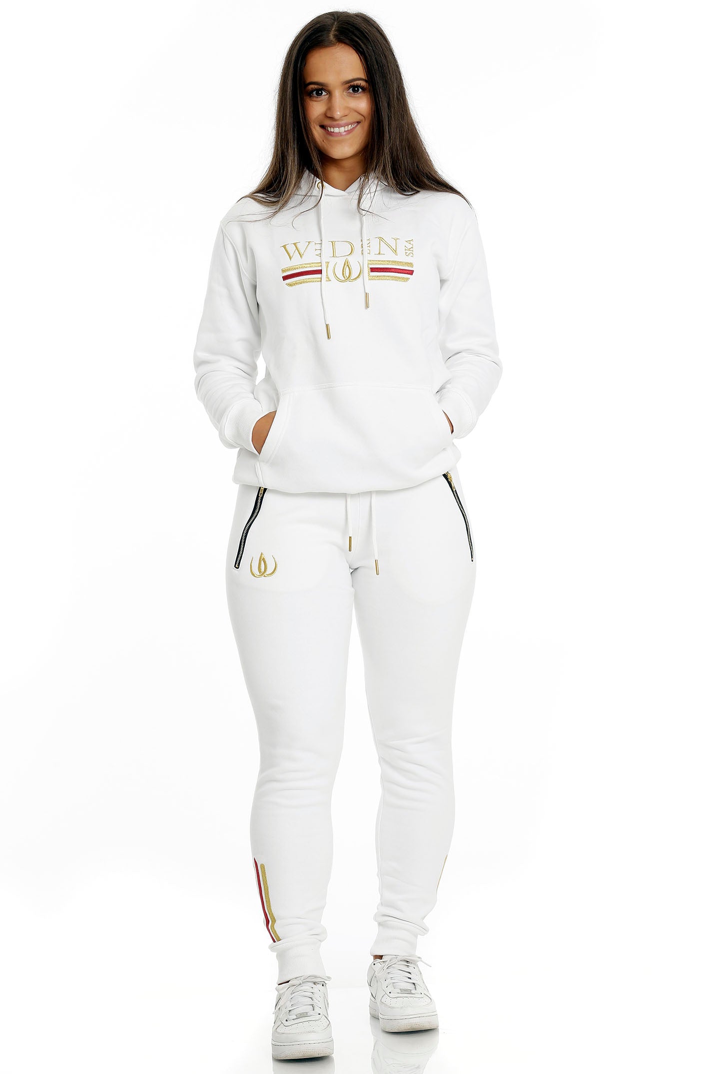 Stående i vita joggers med guldlogga och vit huvtröja med guldbroderi, till vita sneakers.