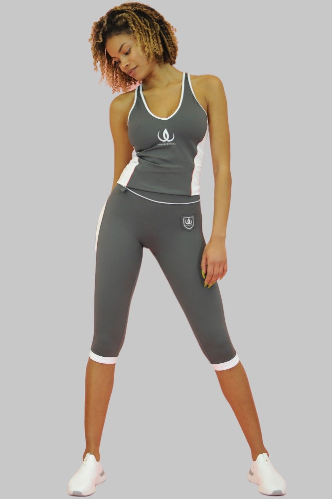 Le legging capri gris porte un logo bouclier blanc sur la cuisse et des bords blancs en bas. Le modèle porte un débardeur gris.