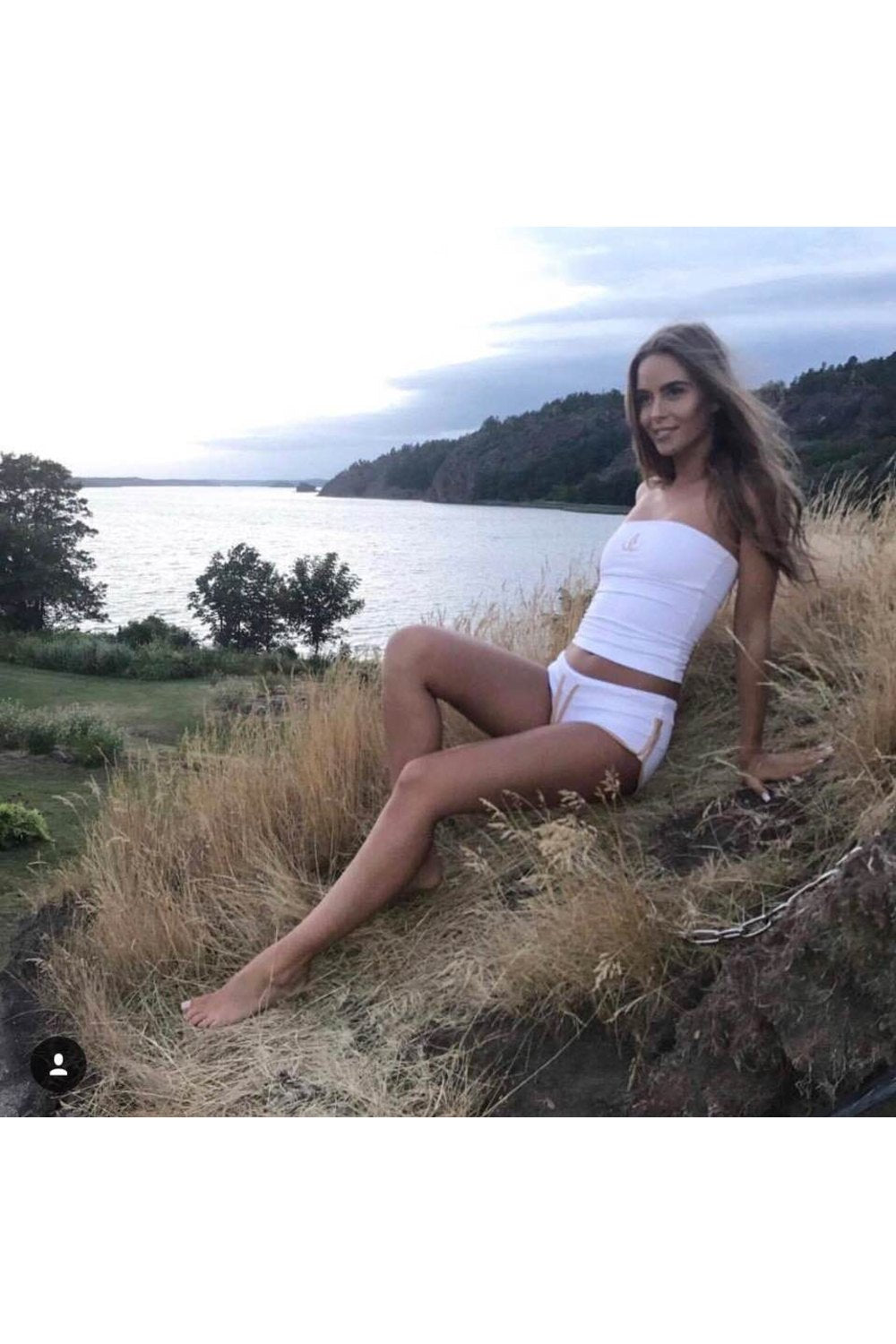 Das Model lehnt sich im Gras am Wasser zurück, in weißen Shorts mit Goldkante und weißem Bandeau-Top.