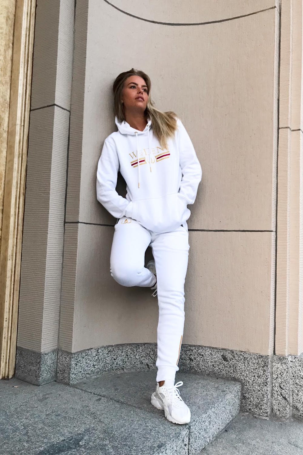 An der Hauswand steht das Model im weißen Hoodie und weißen Joggers, dazu weiße Sneaker.