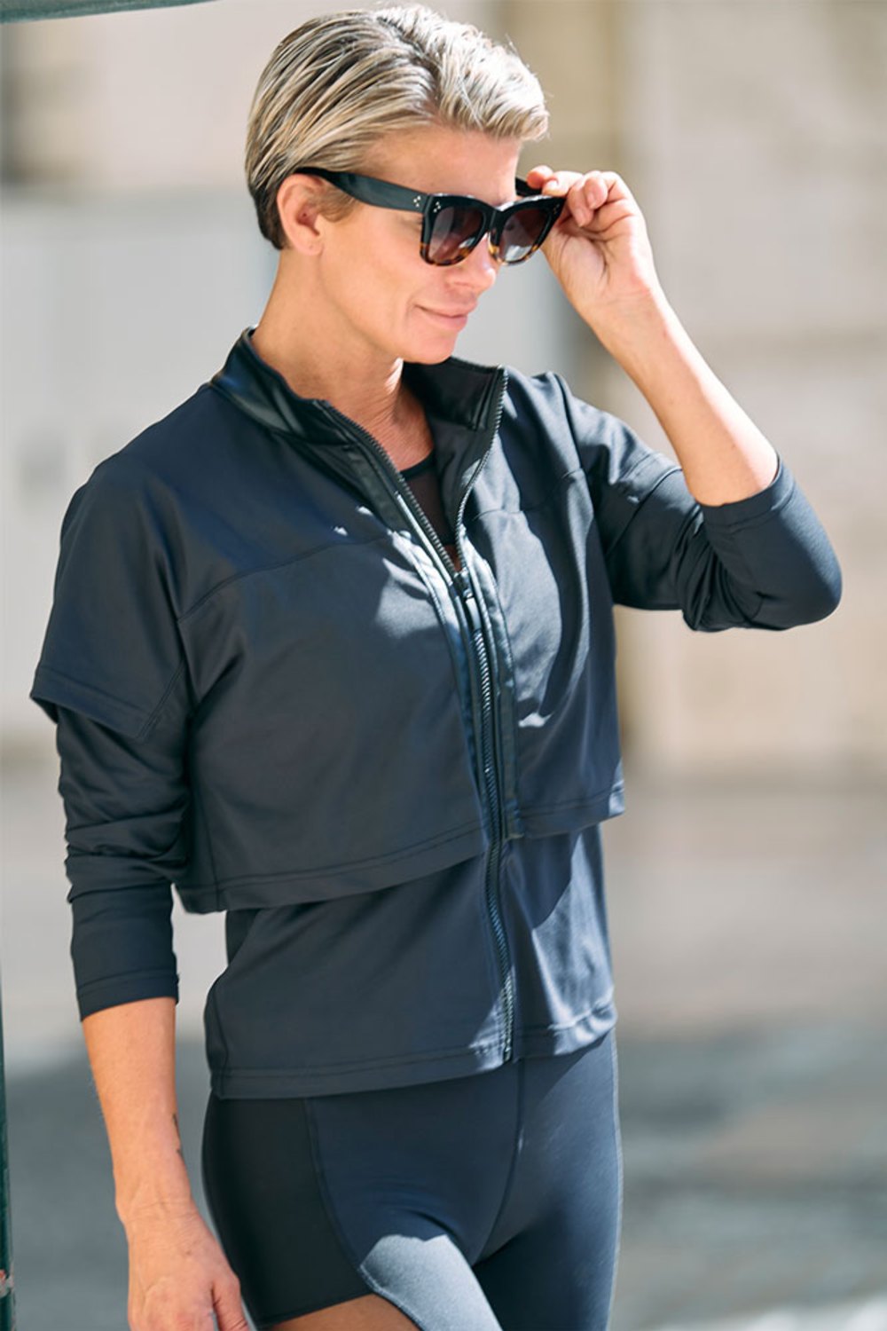 Halb geöffneter Reißverschluss an der schwarzen Jacke, während das Model die Sonnenbrille anhebt.