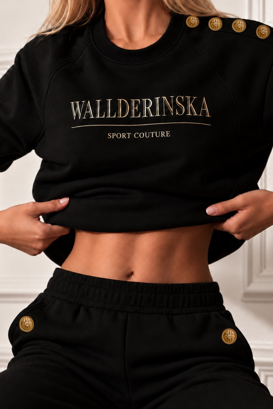 Edge Gold Button Pants i svart med guldknappar vid fickorna, och tröjan lyft över midjan.