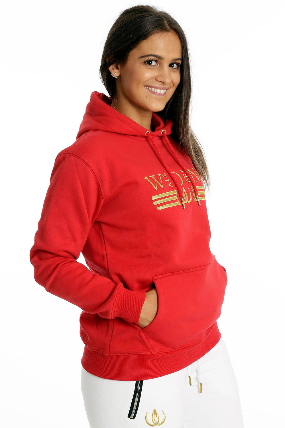 Die Hände ruhen in der Bauchtasche des roten Hoodies. Darunter zeigen sich weiße Jogger mit goldfarbenem Reißverschluss.