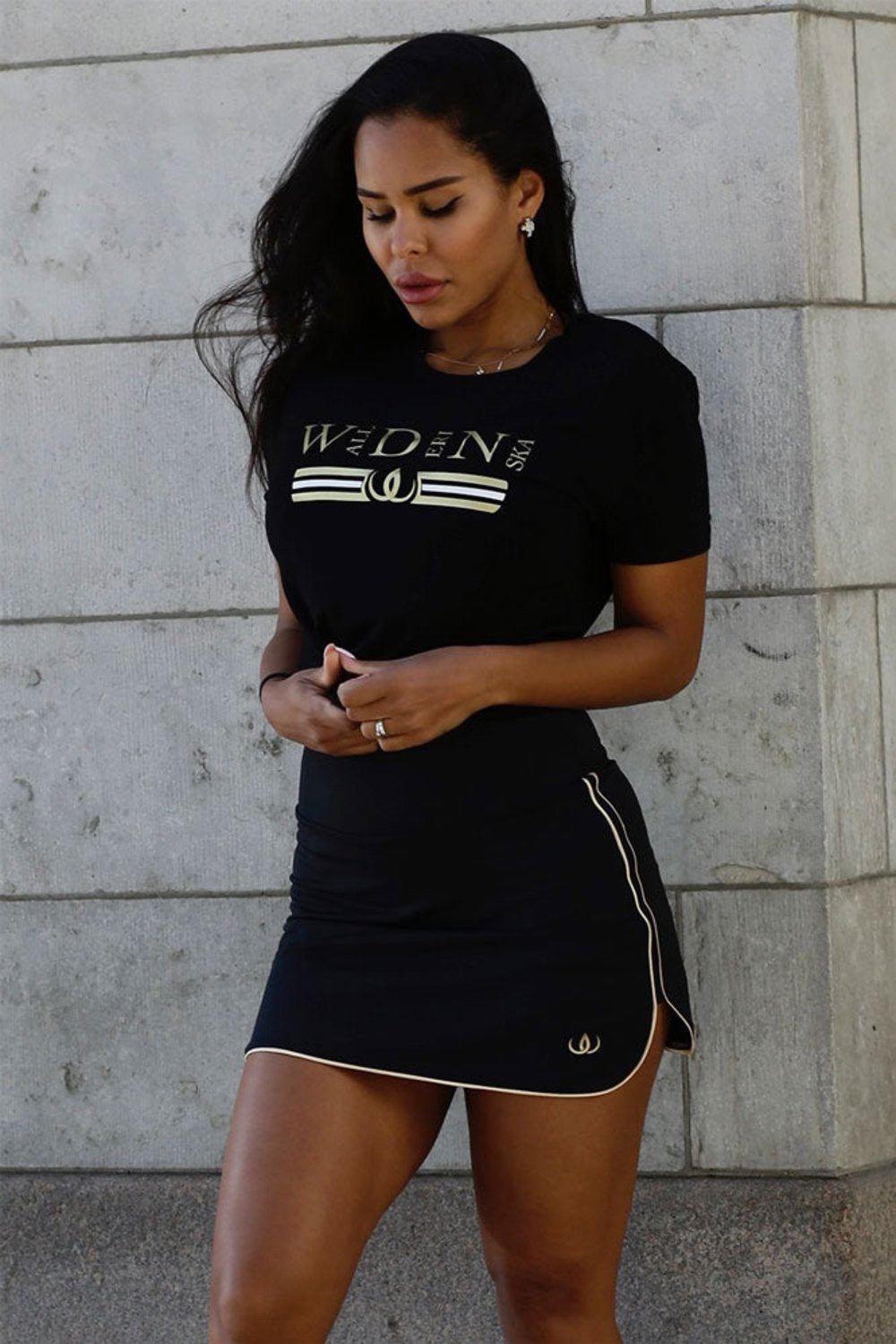 Das Model steht an einer hellen Wand im schwarzen Rock und einem schwarzen T-Shirt mit Golddruck.