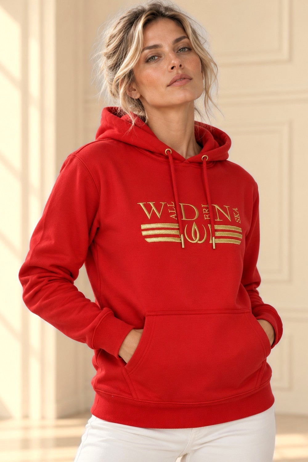 Händerna vilar i magfickan på den röda Deluxe Hoodie med guldbroderad logga.