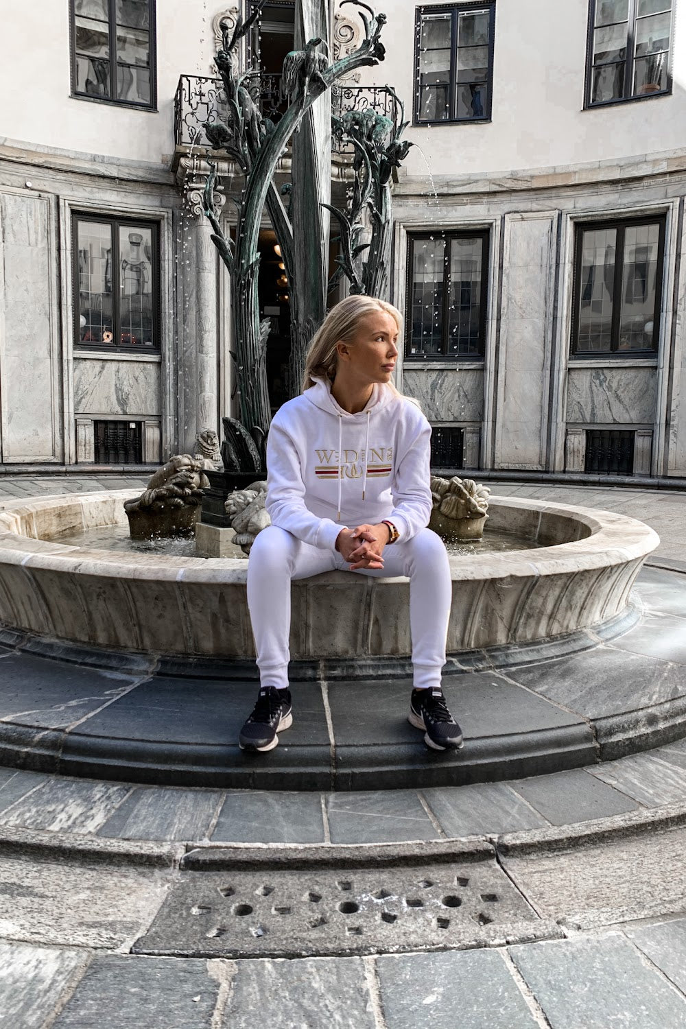 Auf dem Brunnenrand sitzt das Model im weißen Hoodie, mit einer Statue im Hintergrund.