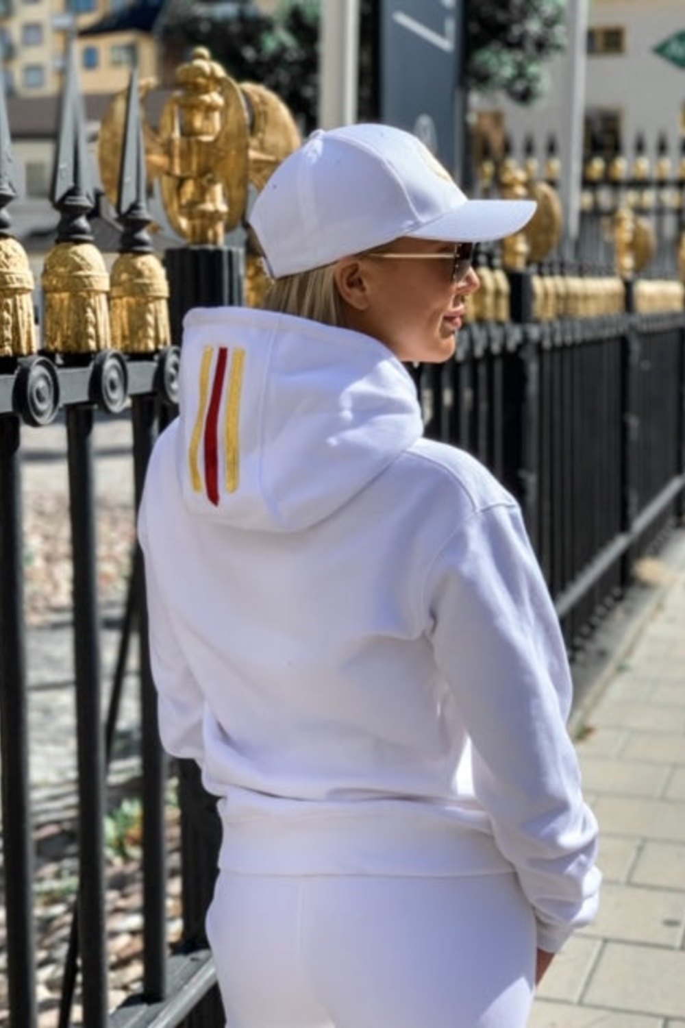 Gold und ein roter Streifen auf der Kapuze sind zu sehen, während das Model einem schwarzen Zaun zugewandt steht.