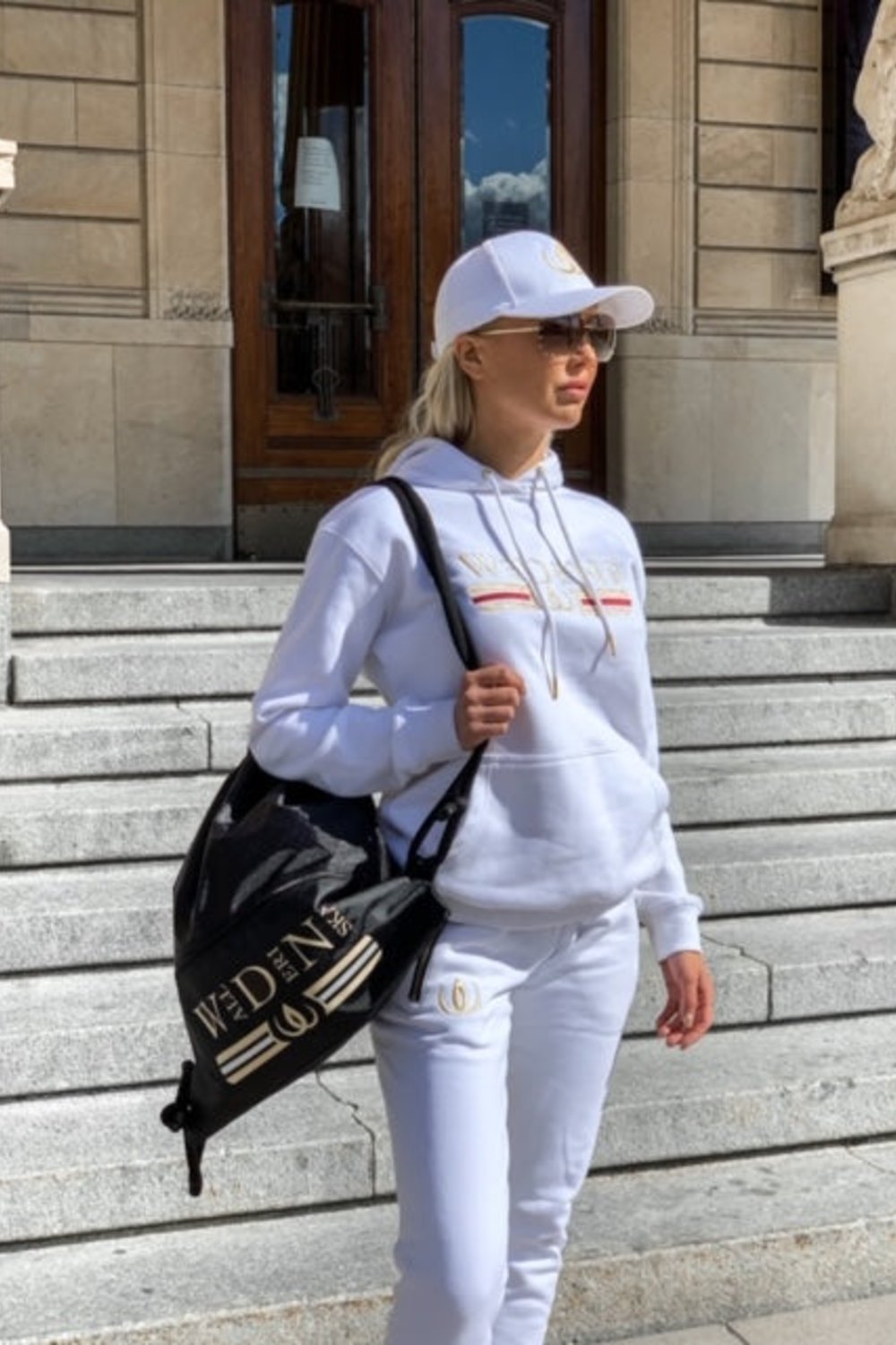 Auf der Treppe vor einem Gebäude wird der weiße Hoodie getragen, mit Cap und dem Deluxe Rucksack Schwarz über der Schulter.