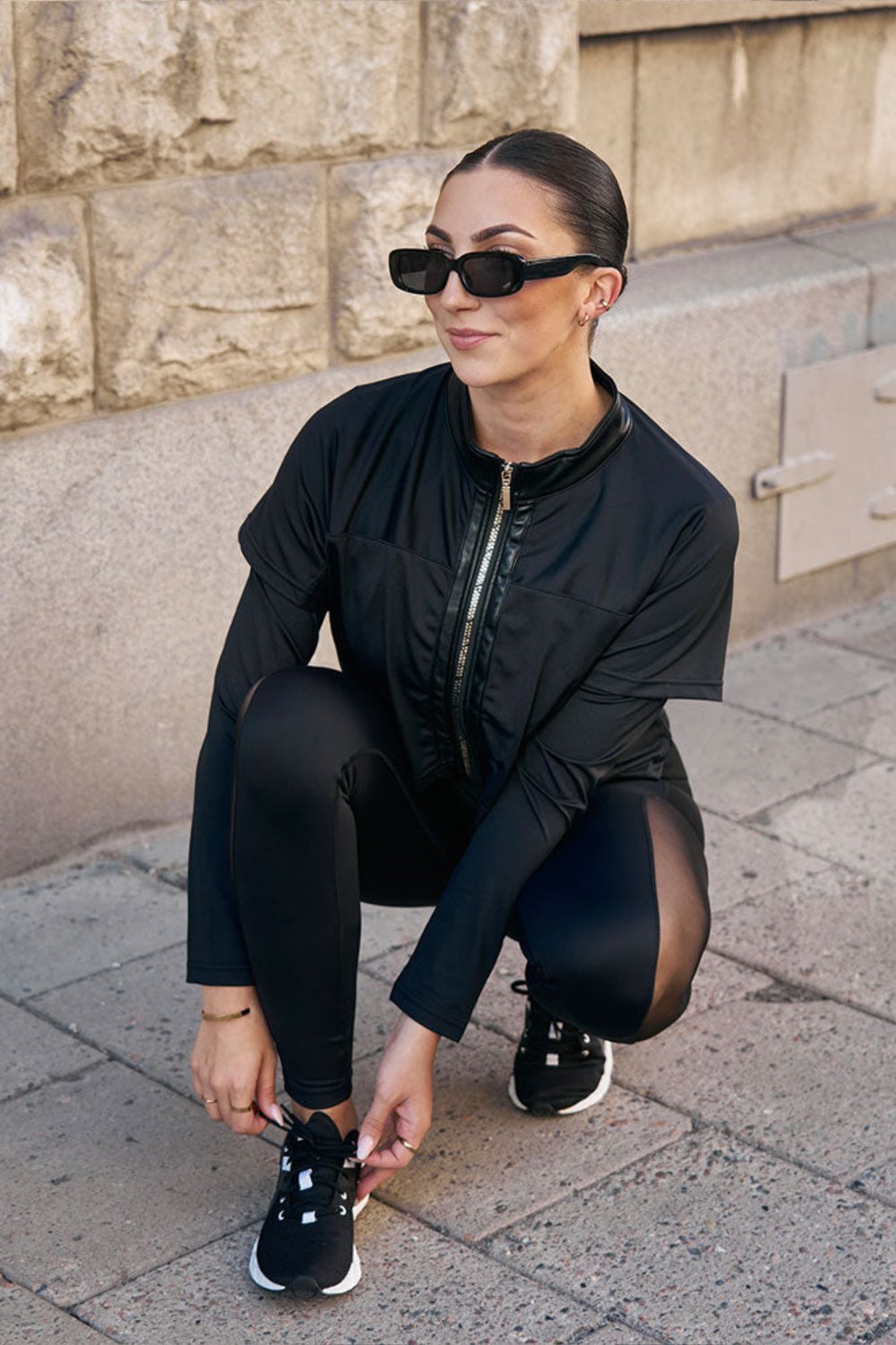 Am Gehweg bindet das Model in der Hocke den Schuh, in der schwarzen Jacke und schwarzer Leggings.