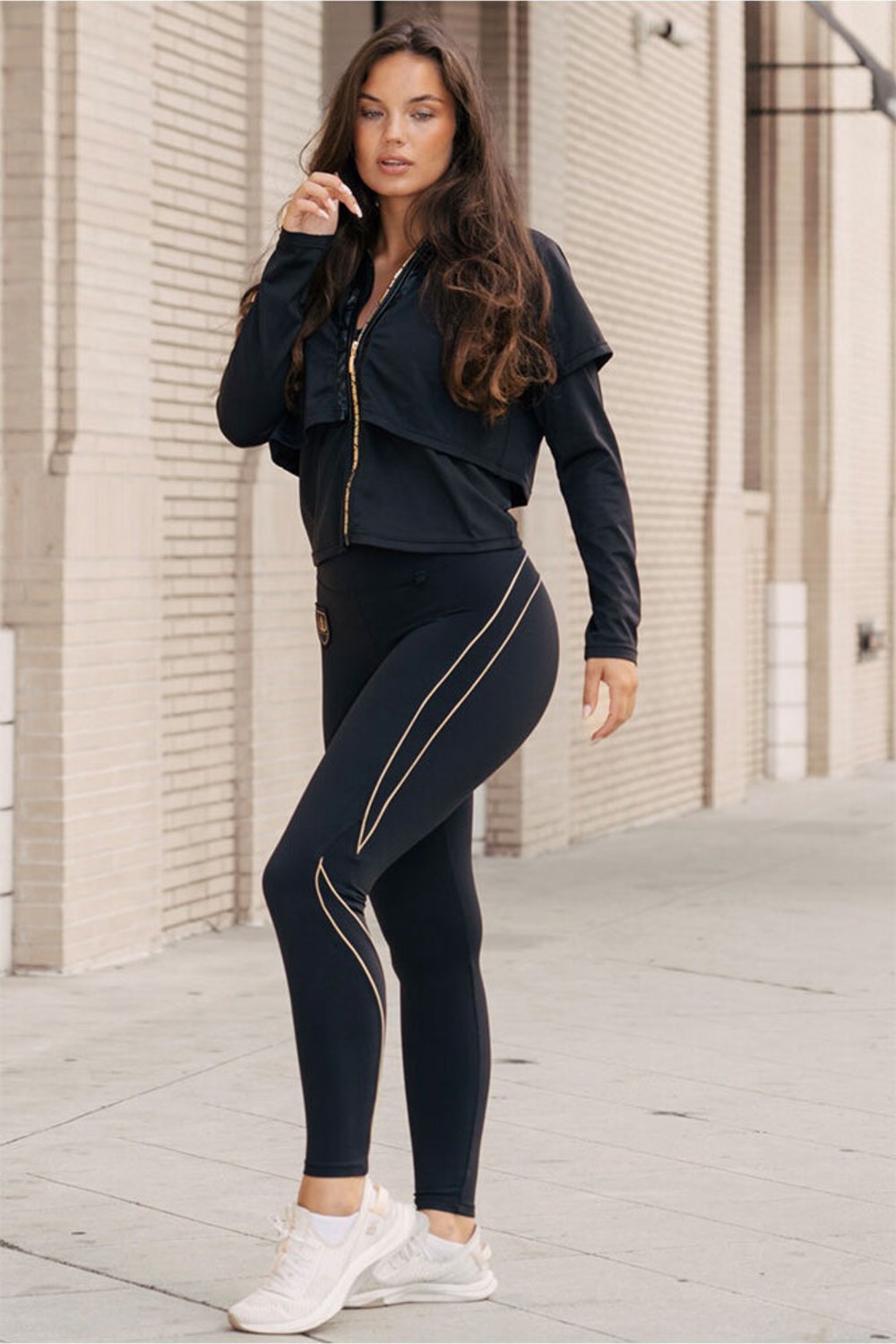 Vor der Ziegelwand steht das Model in der Jacke und schwarzen Leggings mit Goldlinien.
