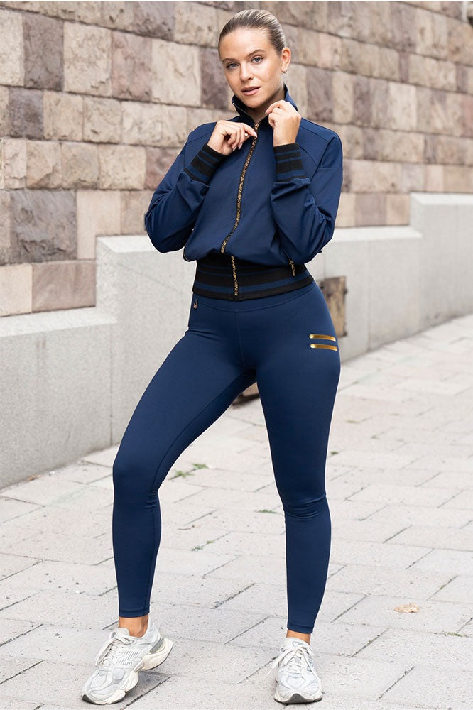 Accanto al muro di pietra, la modella indossa i leggings navy e una giacca con zip color oro.