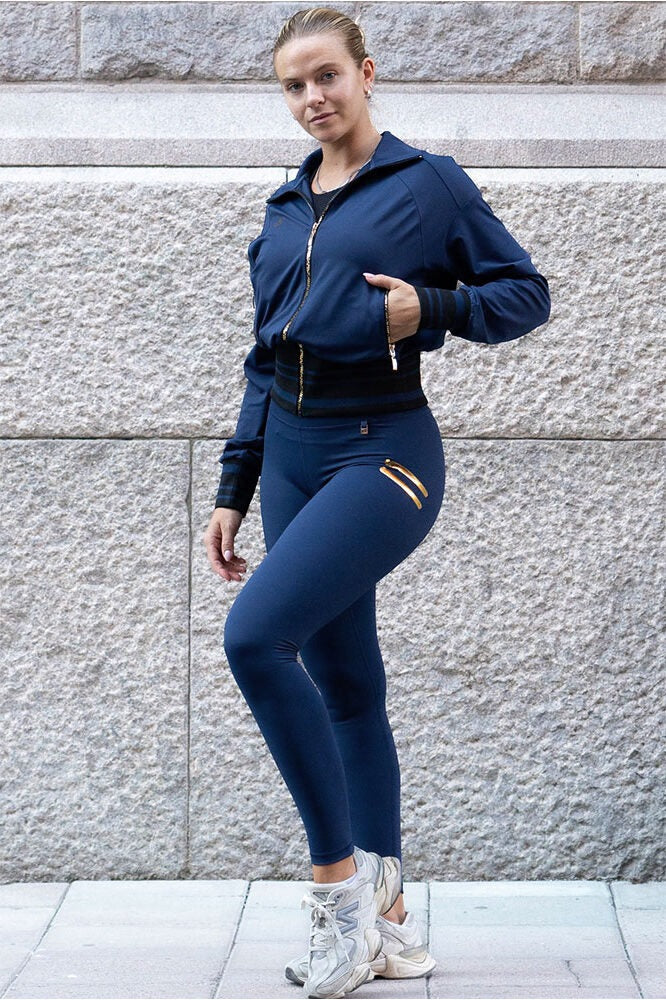 Contro la parete di granito, i leggings navy con una giacca aperta con zip color oro.