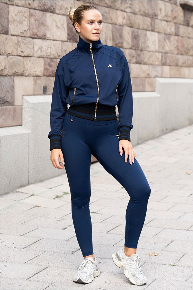 En extérieur, en veste bleu marine à fermetures dorées, legging bleu marine et sneakers blanches.